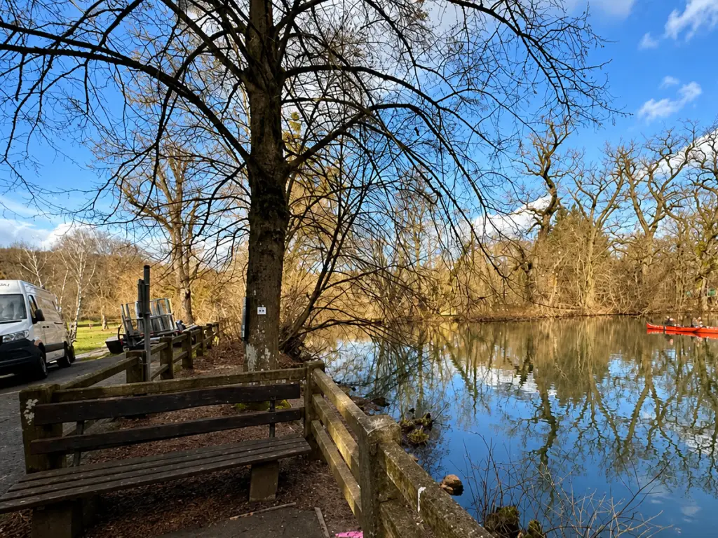 Parkbank am Ufer eines ruhigen Sees mit kahlen Bäumen, Spiegelung im Wasser und rotem Kanu im Hintergrund.