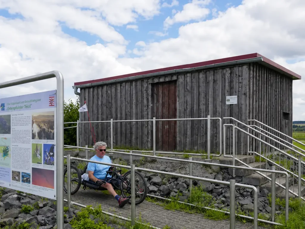 Person fährt mit einem Liegerad auf einer barrierefreien Rampe vor einem Holzgebäude neben einer Infotafel.