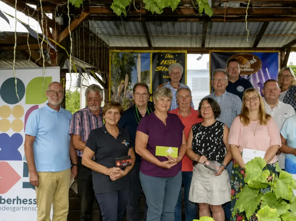 Gruppe von Erwachsenen steht unter einem überdachten Holzpavillon und hält Broschüren oder Karten in den Händen.