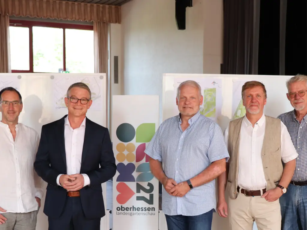 Fünf Männer stehen in einem Innenraum vor Präsentationstafeln und einem Logo und posieren für ein Gruppenfoto.