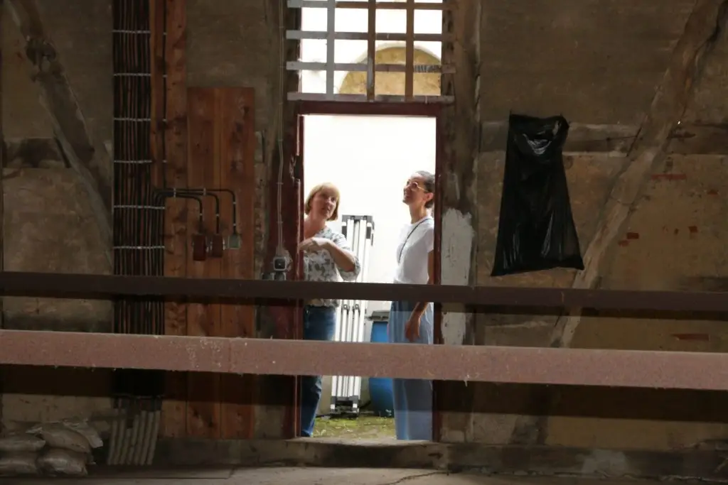 Zwei Frauen stehen in einer offenen Tür eines historischen Gebäudes und schauen sich den Innenraum einer Baustelle mit freigelegten Wänden und Installationen an.