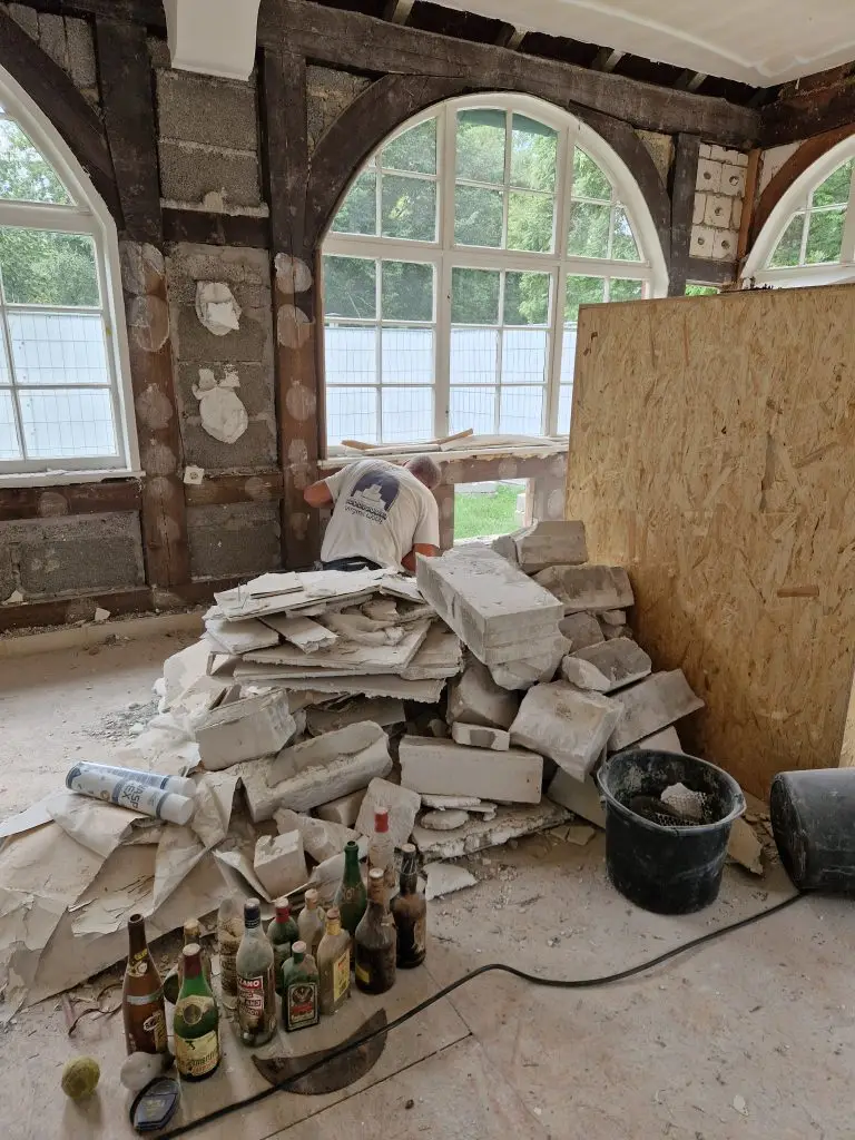 Ein Bauarbeiter kniet vor einem großen Haufen aus Bauschutt und entfernt Material in einem Raum mit freigelegtem Fachwerk und hohen Rundbogenfenstern.