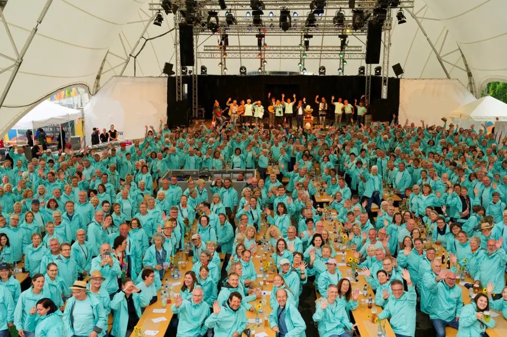 Große Gruppe von Ehrenamtlichen in einheitlichen T-Shirts sitzt in einem Festzelt und blickt zur Bühne.