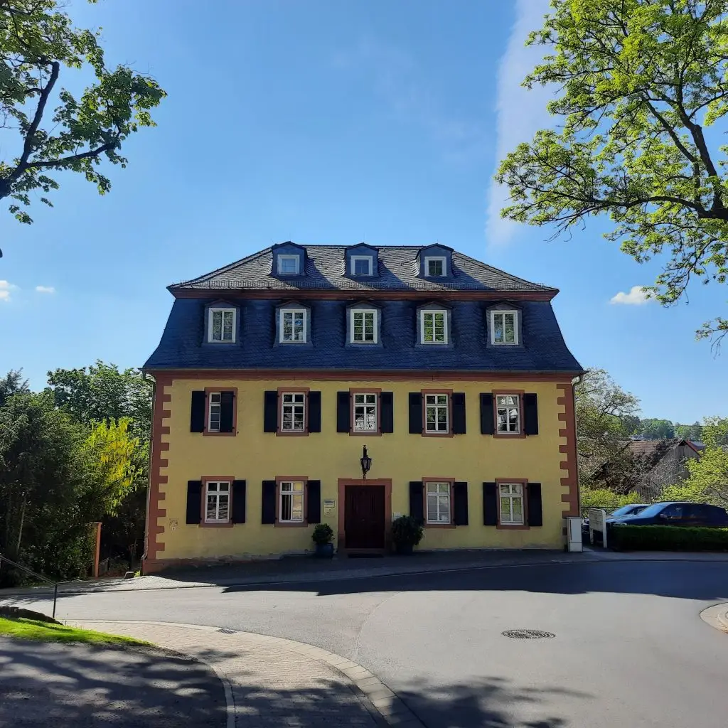 Ein dreistöckiges historisches Gebäude mit gelber Fassade, dunklen Fensterläden und Mansarddach steht an einer Straße, umgeben von Bäumen.