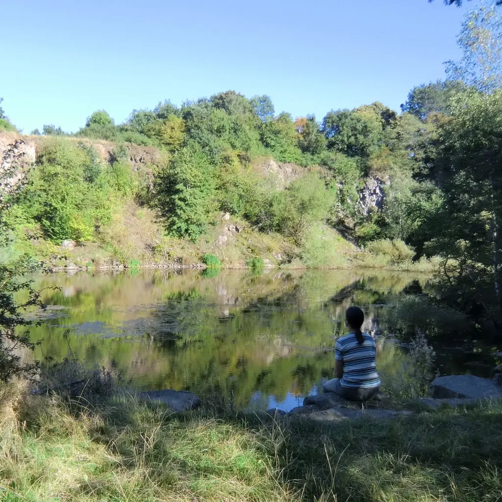Eine Person sitzt am Rand eines Sees und schaut auf das ruhige Wasser, das von Bäumen und Felsen umgeben ist.