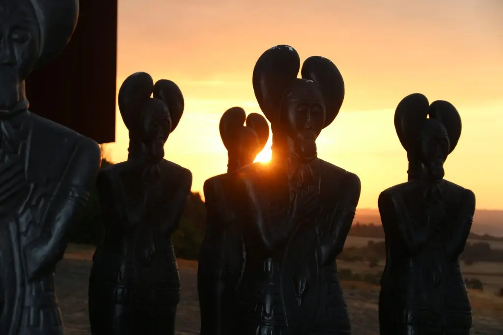 Mehrere dunkle, stilisierte Figuren stehen vor einem Sonnenuntergang, während die Sonne zwischen ihnen hindurchscheint.