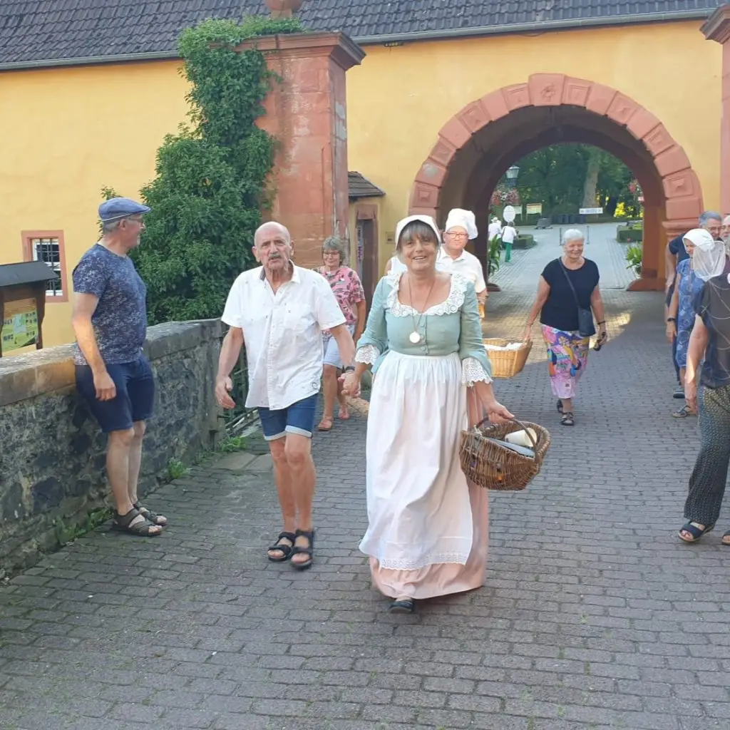 Mehrere Menschen, darunter eine Frau in historischer Kleidung mit Korb, gehen über eine Steinbrücke auf ein Schlosstor zu.