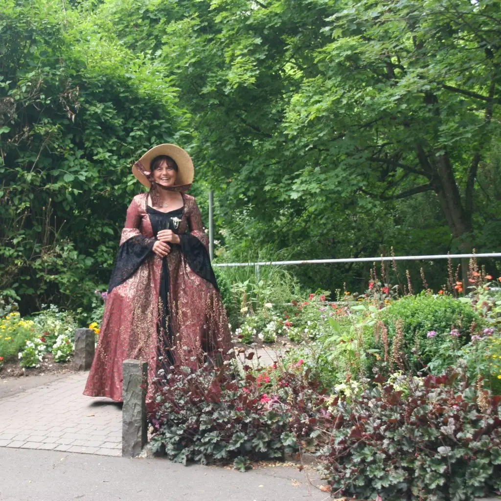 Eine Frau in historischer Kleidung mit Hut steht auf einem Weg in einem Garten mit Blumenbeeten und Bäumen.