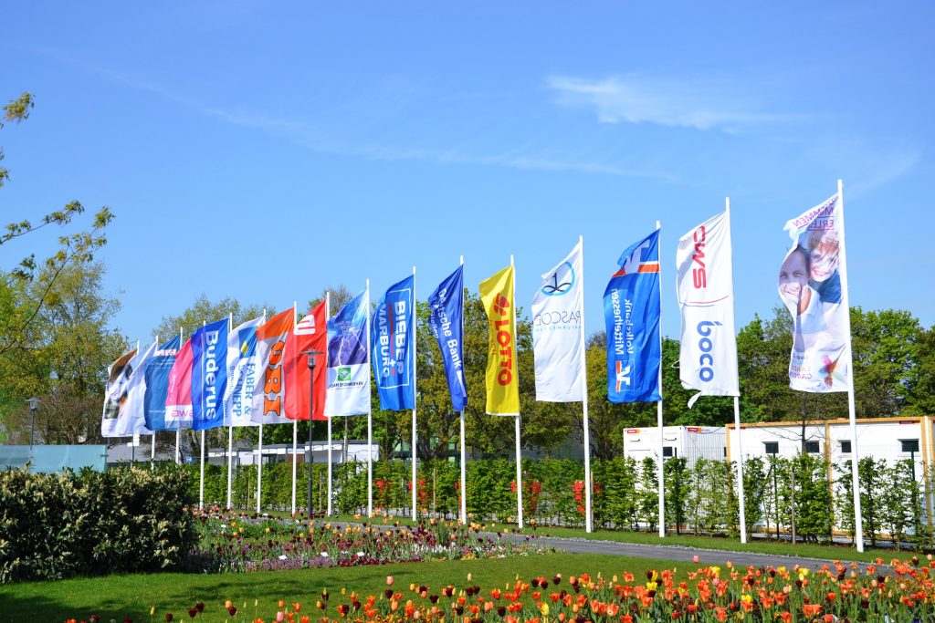 Mehrere Fahnen von Unternehmen und Organisationen wehen entlang einer blühenden Parkanlage vor blauem Himmel.