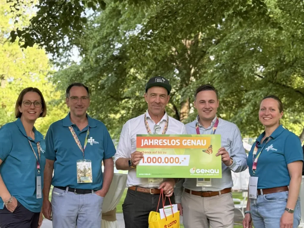 Fünf Personen stehen bei einer Veranstaltung im Freien und präsentieren ein „Jahreslos Genau“ mit Gewinnsumme.