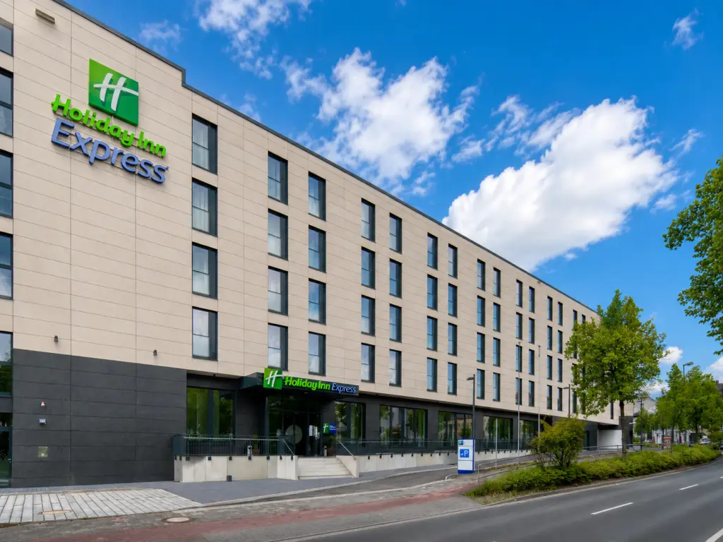 Außenansicht des Holiday Inn Express Fulda mit modernem Hotelgebäude, begrünten Straßenbereichen und blauem Himmel.