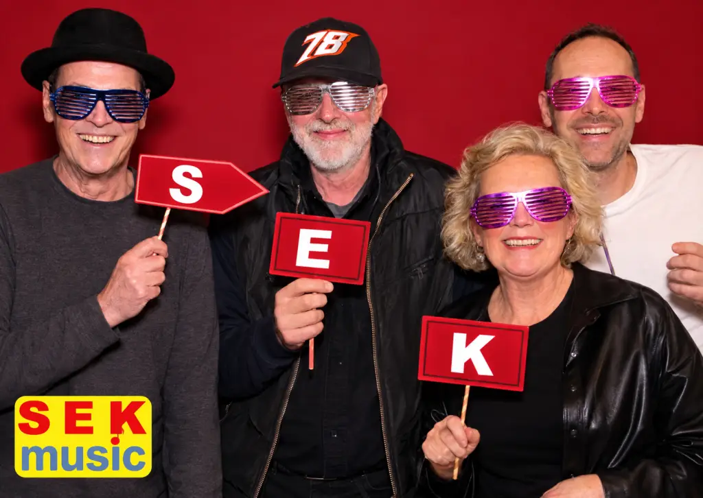 Vier Mitglieder der Band SEK Music mit bunten Partybrillen und roten Schildern mit den Buchstaben S, E und K vor rotem Hintergrund.