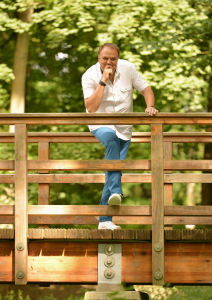 Ein Musiker lehnt auf einer Holzbrücke in einem grünen Wald und blickt in die Kamera.