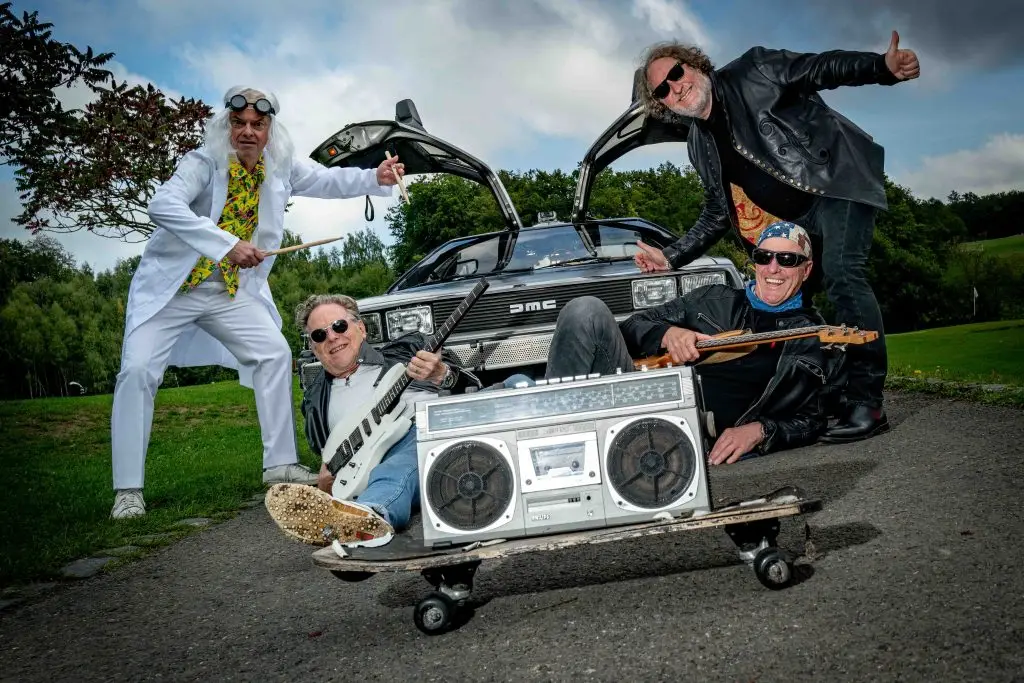 Vier Mitglieder von einer Band posieren mit Gitarren und Ghettoblaster vor einem DeLorean.