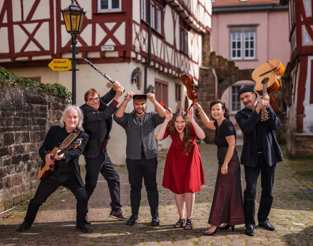 Sechs Musikerinnen und Musiker posieren gut gelaunt in einer gepflasterten Altstadtgasse mit Fachwerkhäusern und halten ihre Instrumente – darunter Gitarre, Klarinette und Geige – spielerisch über ihre Köpfe.