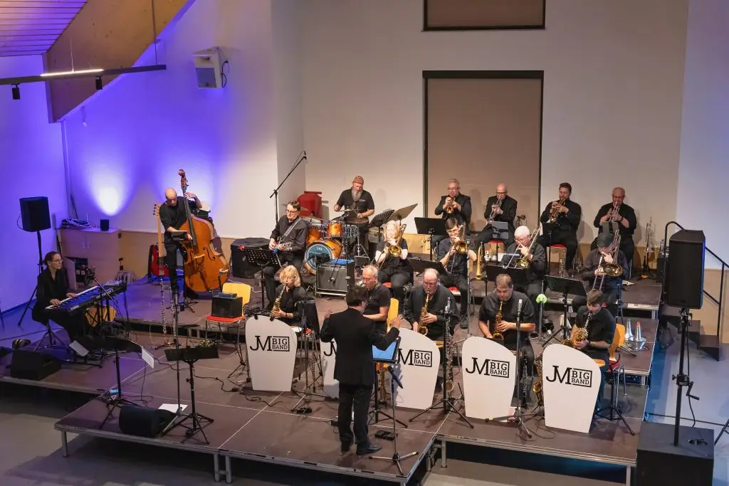 Die JM Big Band spielt unter der Leitung eines Dirigenten auf einer Bühne.