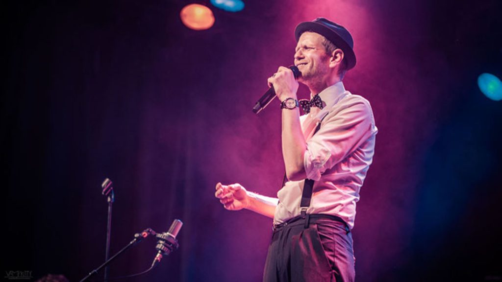 Sänger Kai Podack singt mit Mikrofon auf einer violett beleuchteten Bühne.