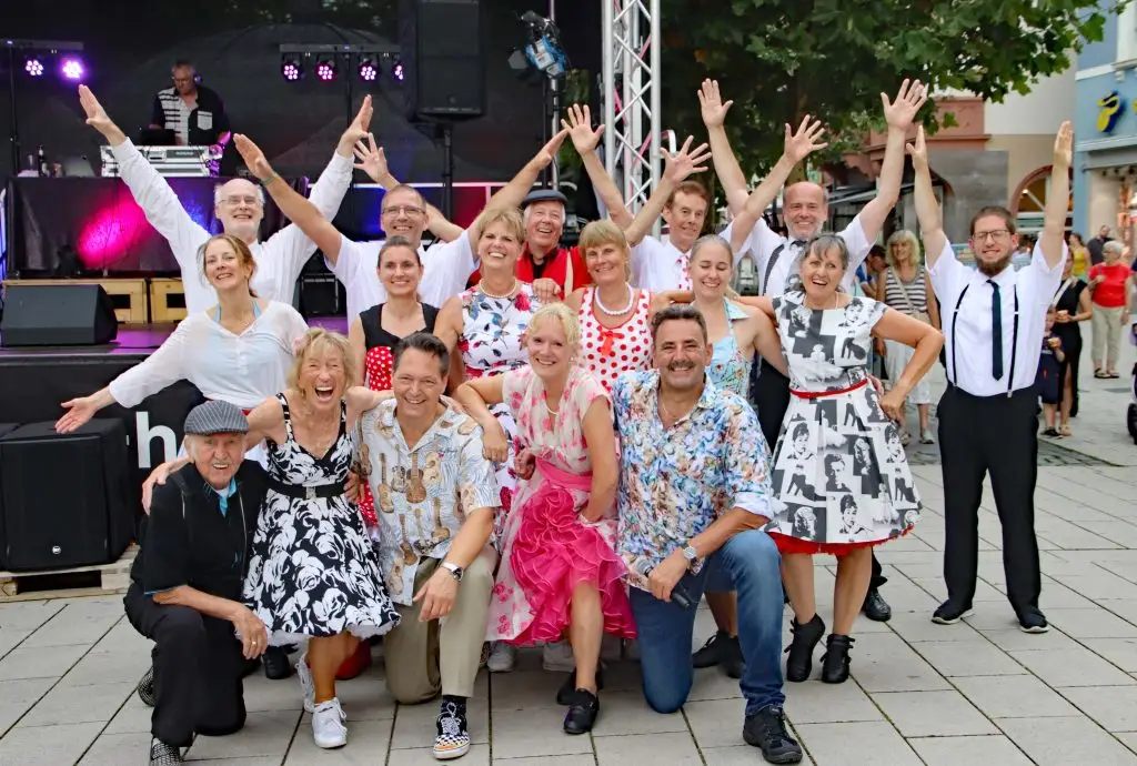 Eine Tanzgruppe posiert gut gelaunt vor einer Bühne und streckt teilweise die Arme in die Höhe.