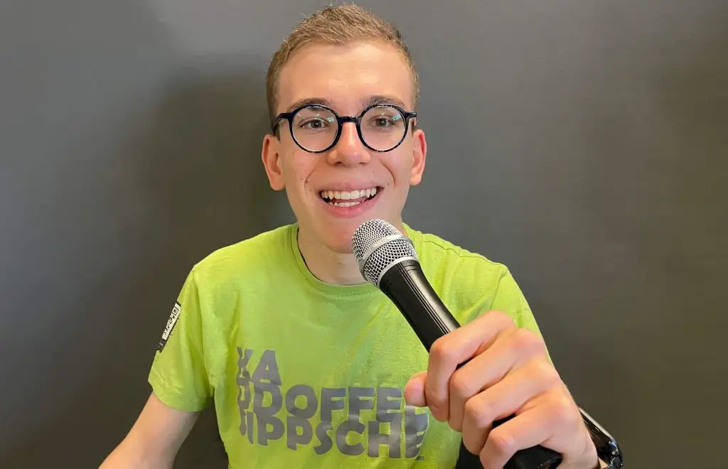 Luis Adam lächelt in die Kamera und hält ein Mikrofon vor sich. Er trägt eine grünes T-Shirt mit Aufschrift und eine runde Brille.
