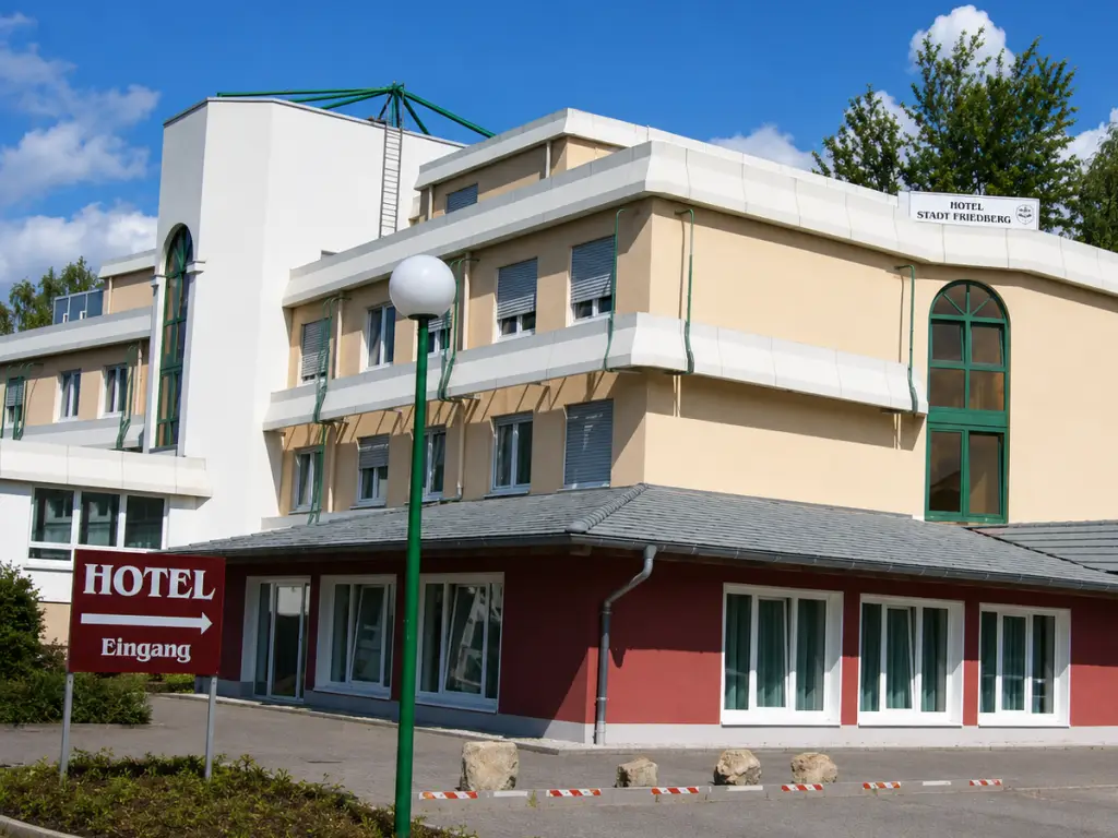 Außenansicht des Hotels Stadt Friedberg mit beigefarbener Fassade, rotem Erdgeschoss und dem Hinweisschild „Hotel Eingang“.