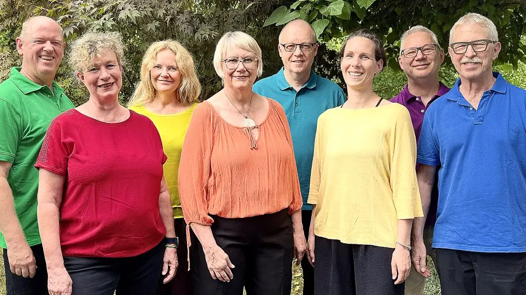 Eine achtköpfige Gruppe steht lächelnd in farbenfroher Kleidung vor einer grünen Baumkulisse.