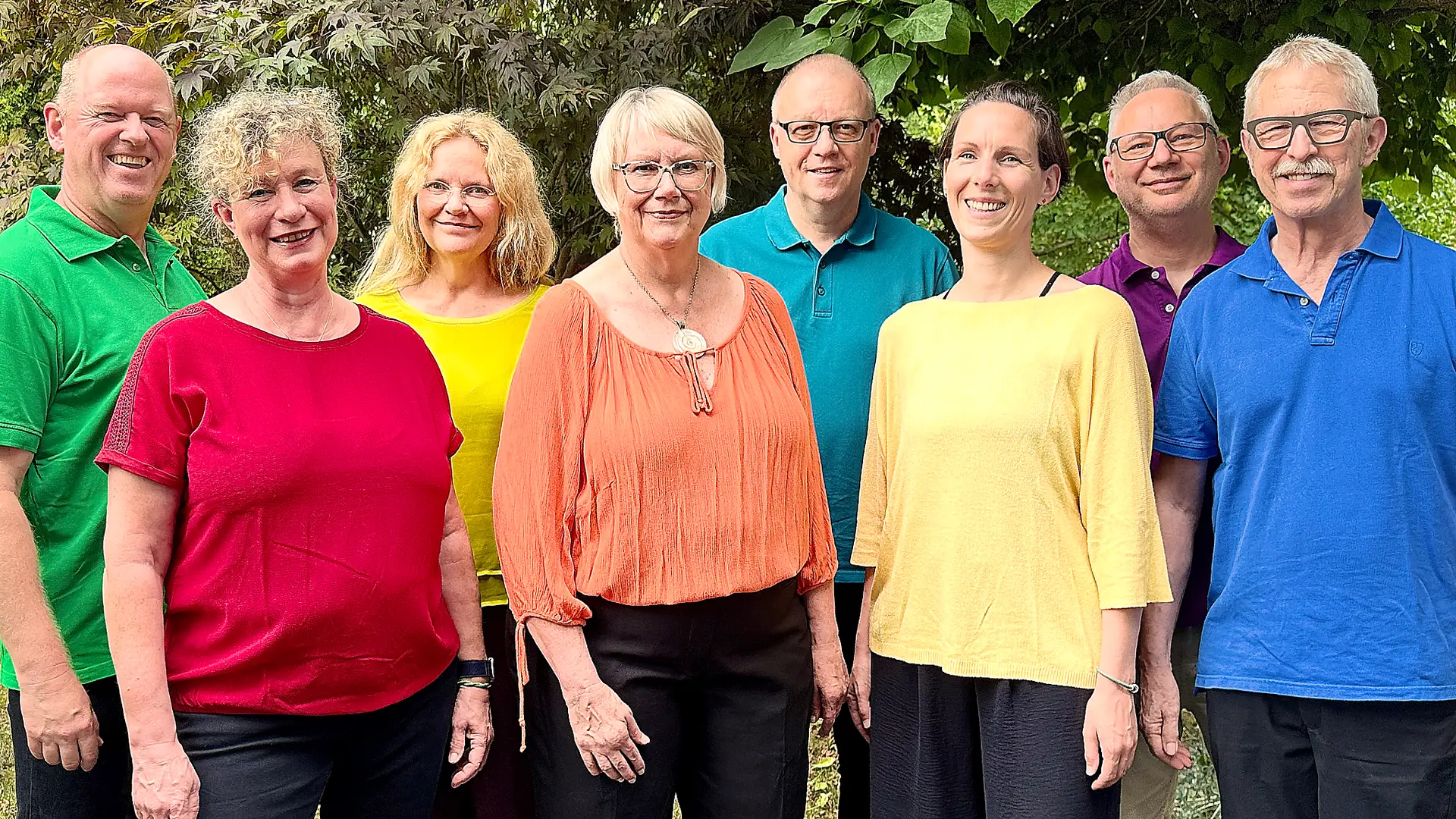 Eine achtköpfige Gruppe steht lächelnd in farbenfroher Kleidung vor einer grünen Baumkulisse.
