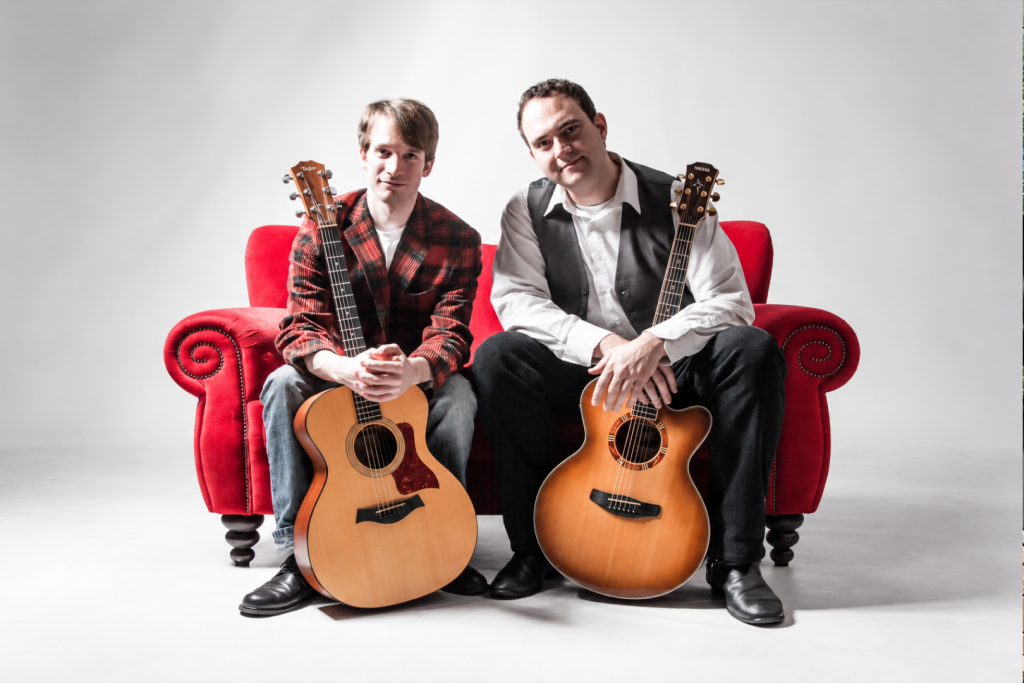 Zwei Musiker sitzen mit Akustikgitarren auf einem roten Sofa.