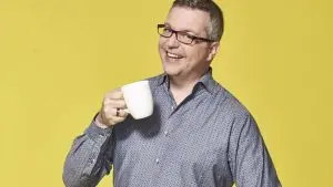 Johannes Scherer steht freudestrahlend vor einem gelben Hintergrund und hält eine Kaffeetasse vor sich.