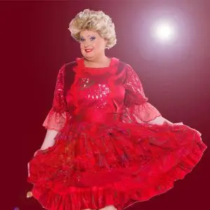 Olga Orange lächelt in die Kamera und präsentiert ein weit ausgestelltes, leuchtend rotes Rüschenkleid. Mit blonder Hochsteckfrisur, auffälligem Make-up und funkelndem Bühnenoutfit strahlt sie eine glamouröse Show-Atmosphäre aus.