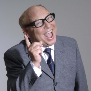 Hans-Joachim Heist schaut mit erhobenem, ermahnendem Finger und lachen in die Kamera. Er trägt einen grau melierten Anzug, sowie eine markante Brille mit breitem schwarzem Rahmen.