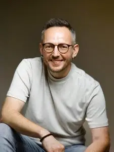 Porträt des Autors Daniel Holbe mit Brille und hellem T-Shirt vor neutralem Hintergrund.