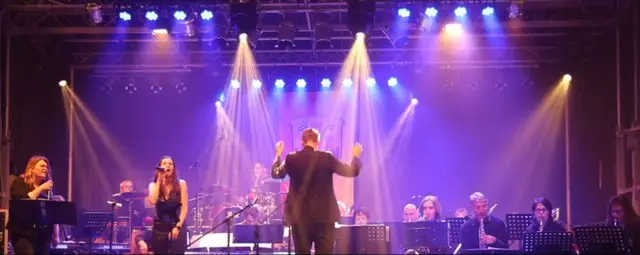 Eine Band mit Sängerinnen, Musikern und Dirigent tritt auf einer farbig beleuchteten Bühne auf.