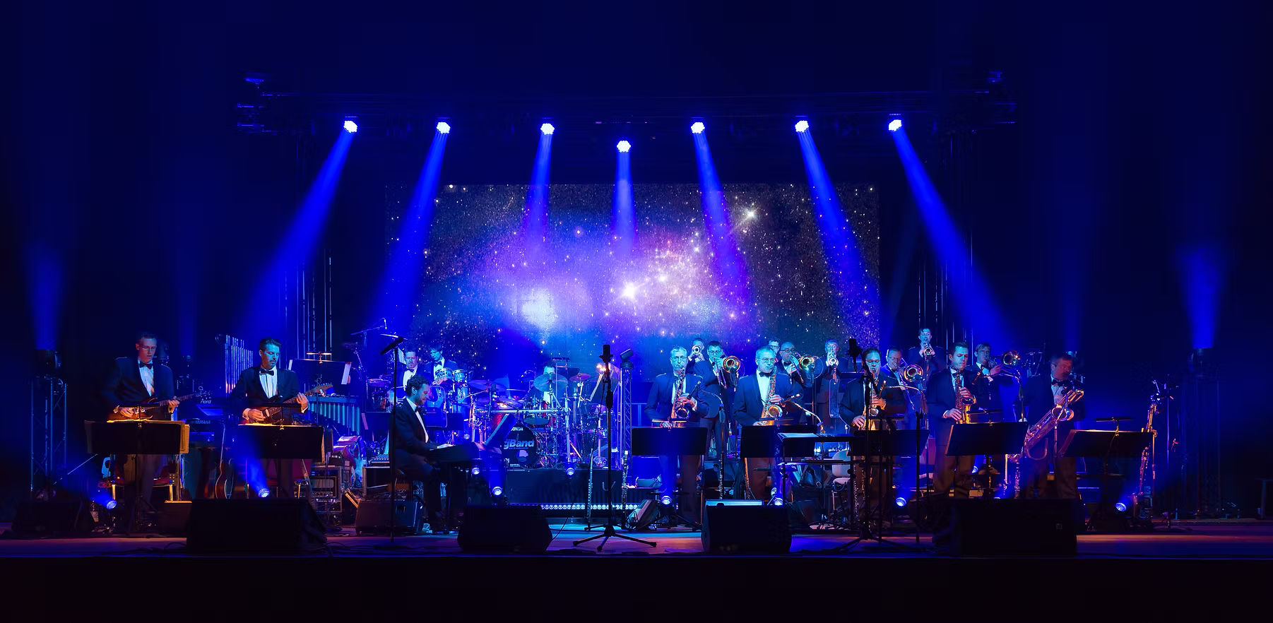 Eine Big Band spielt mit Saxofonen, Posaunen, Trompeten, Schlagzeug und weiteren Instrumenten auf einer blau beleuchteten Bühne.
