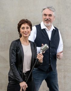 Ein musikalisches Duo posiert lächelnd mit einem Retro-Mikrofon.