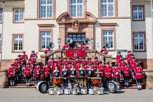 Große Musikgruppe in roten Uniformen posiert mit Blas- und Schlaginstrumenten vor einem historischen Gebäude.