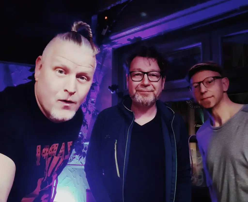 Die drei Mitglieder der Band Green Rock posieren unter violett-blauer Beleuchtung für die Kamera.