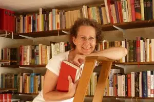 Astrid Hohlbein lächelt mit einem roten Buch vor einem Bücherregal.