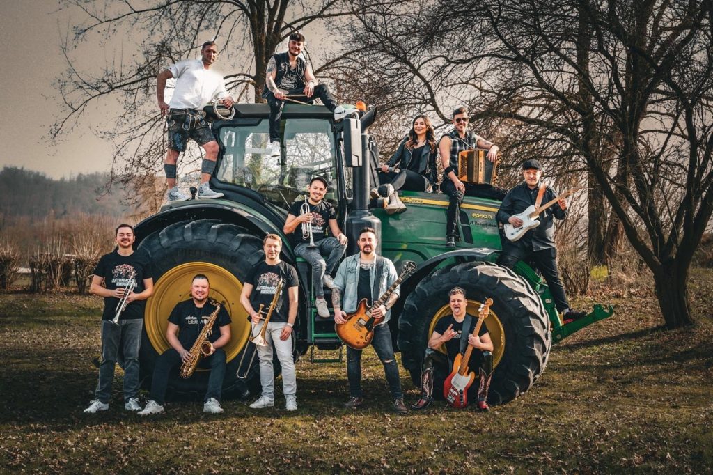 Eine große Band posiert mit verschiedenen Instrumenten rund um einen grünen Traktor.
