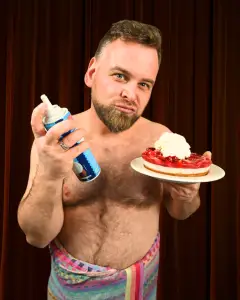 Musicalstar André Haedicke posiert mit Sahnespray und einer Torte vor einem roten Bühnenvorhang.