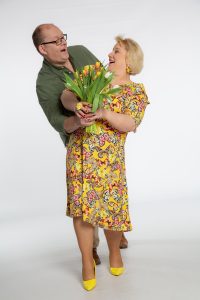 Ein Künstler-Duo posiert lachend mit einem bunten Tulpenstrauß vor weißem Hintergrund.