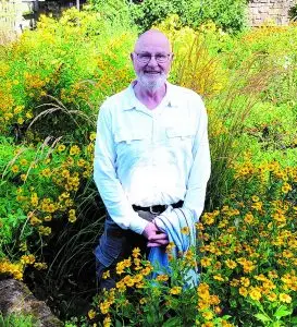 Ein lächelnder Autor steht inmitten eines üppig blühenden Gartens mit gelben Blumen.