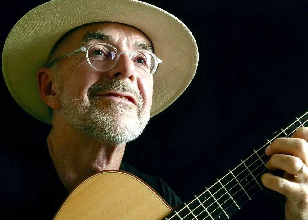 Ein Musiker mit Hut und Brille spielt eine akustische Gitarre vor dunklem Hintergrund.