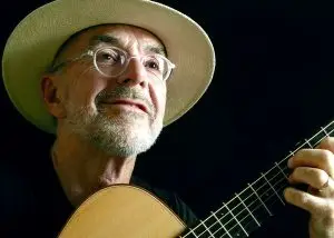 Ein Musiker mit Hut und Brille spielt eine akustische Gitarre vor dunklem Hintergrund.