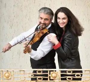 Das Duo Bravo posiert lächelnd mit einer Violine vor einer hellen Wand.