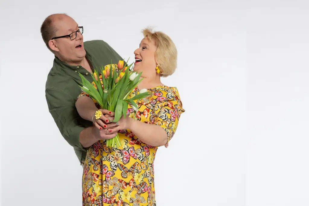 Ein Künstler-Duo posiert lachend mit einem bunten Tulpenstrauß vor weißem Hintergrund.