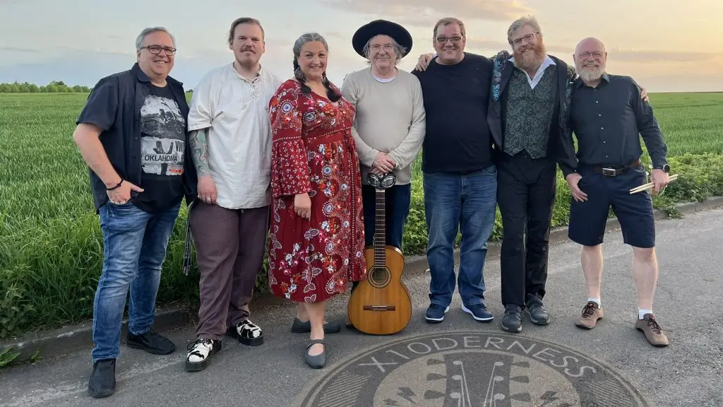 Eine siebenköpfige Musikgruppe posiert mit Gitarre und Drumsticks am Rand eines Feldes.