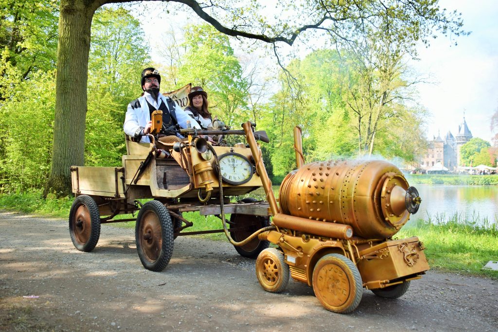 Zwei Personen in Steampunk-Kostümen fahren mit einem fantasievollen, goldfarbenen Gefährt durch einen Park.