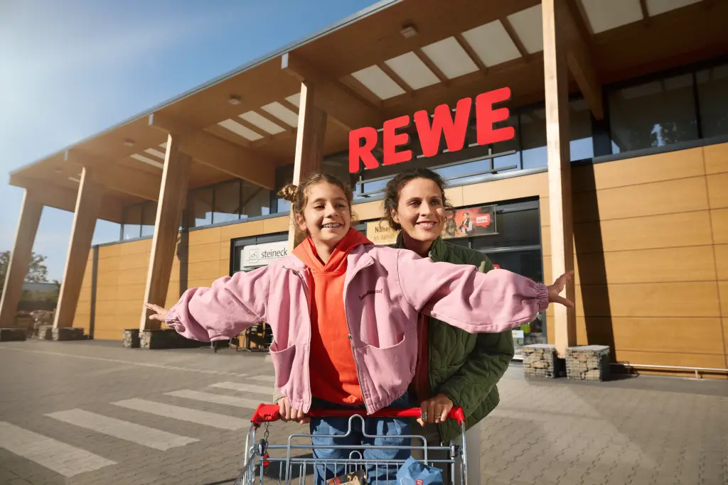 Mutter und Tochter stehen mit einem Einkaufswagen vor einem REWE-Markt.