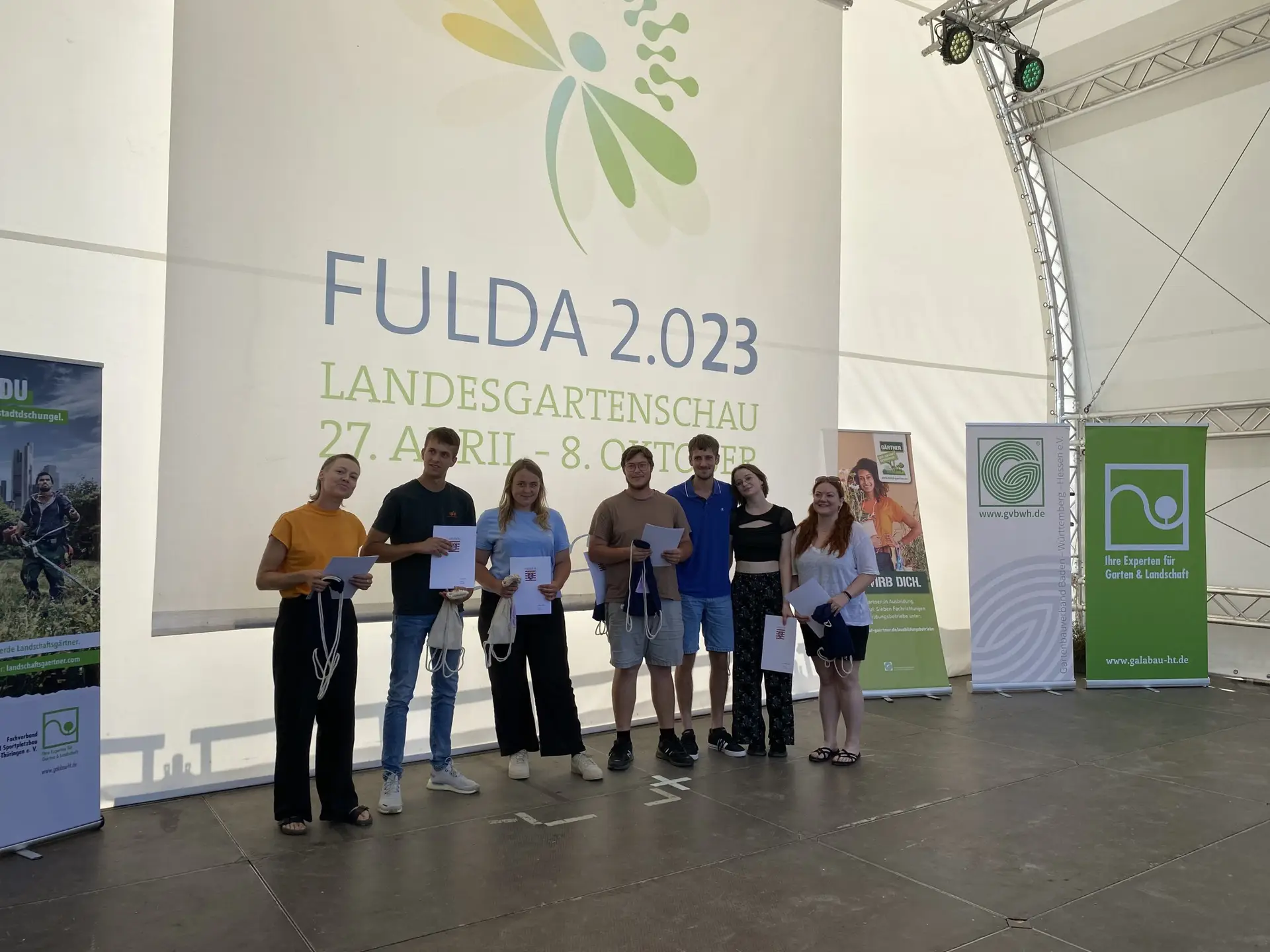 Sieben junge Erwachsene stehen mit Urkunden und Stoffbeuteln auf einer Bühne der Landesgartenschau Fulda 2023.