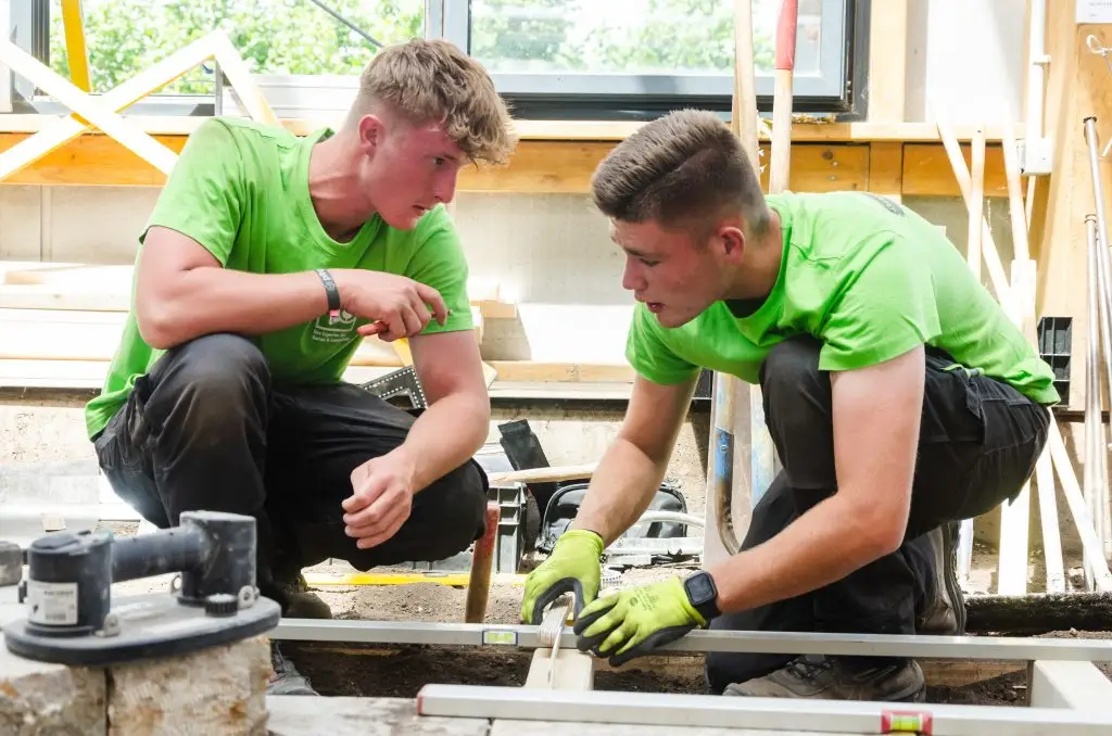 Zwei junge Handwerker arbeiten gemeinsam mit Wasserwaage und Holz an einer Konstruktion.